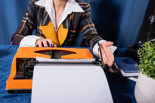 Corte vista de newswoman elegante perto de máquina de escrever vintage com papel em branco no fundo azul — Fotografia de Stock