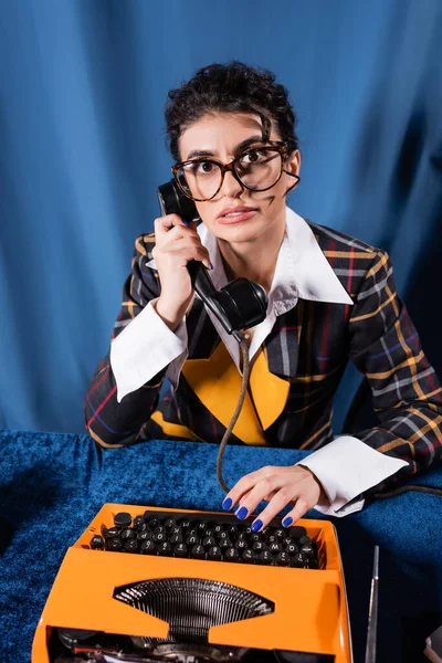 Irritierte Nachrichtensprecherin im Vintage-Stil blickt in die Kamera, während sie in der Nähe einer Schreibmaschine auf blauem Hintergrund telefoniert — Stockfoto