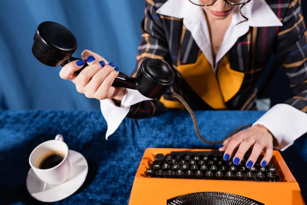 Partial view of stylish newswoman holding handset while typing on vintage typewriter on blue background — Stock Photo