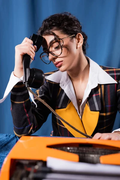 Cansada reportera sentada con el teléfono cerca de la máquina de escribir retro sobre fondo azul - foto de stock