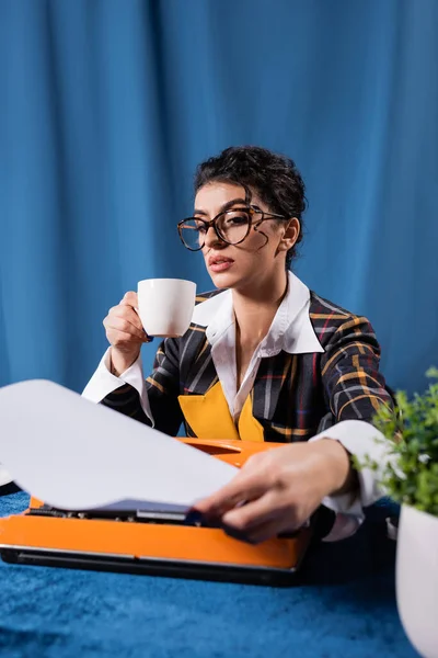 Vintage style newswoman with coffee cup holding empty paper near typewriter on blue background — Stock Photo