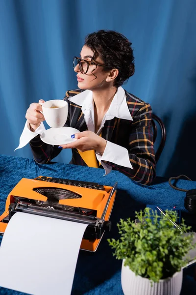 Jornalista elegante sentado com xícara de café perto de máquina de escrever vintage em fundo azul — Fotografia de Stock