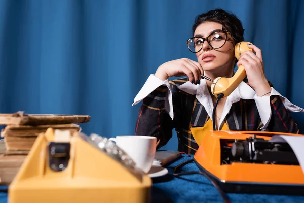 Pensativa reportera hablando por teléfono borroso cerca de la máquina de escribir sobre fondo azul - foto de stock