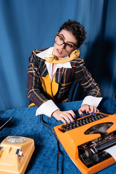 Jornalista em roupas de estilo vintage chamando no telefone enquanto digita no fundo azul — Fotografia de Stock