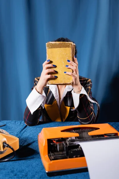 Newswoman obscuring face with book near vintage typewriter on blue background — Stock Photo