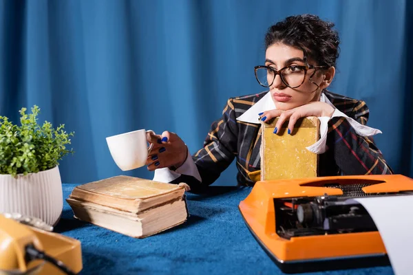 Mujer pensativa sentada con taza de café y libro cerca de máquina de escribir vintage sobre fondo azul - foto de stock