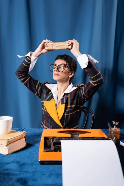 Retro estilo reportero meditando con libro por encima de la cabeza cerca de máquina de escribir sobre fondo azul - foto de stock