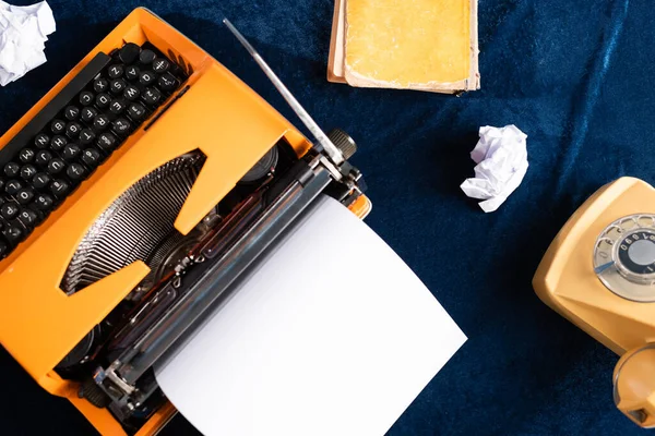 Вид винтажной пишущей машинки с чистой бумагой, телефоном и книгой на голубой скатерти — стоковое фото