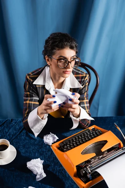 Nachdenkliche Zeitungsfrau mit zerknittertem Papier, die in der Nähe einer alten Schreibmaschine auf blauem Hintergrund wegschaut — Stockfoto