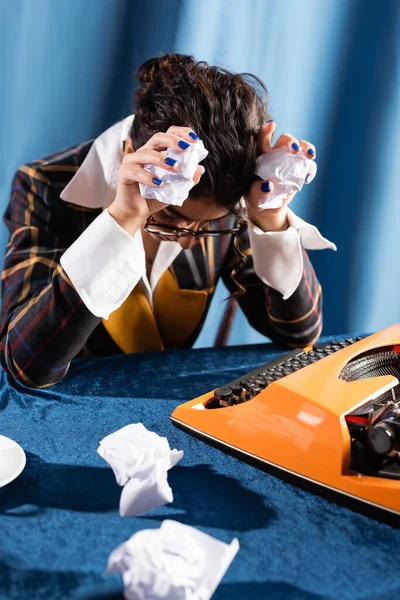 Depressed journalist sitting with bowed head and crumpled paper near vintage typewriter on blue background — Stock Photo