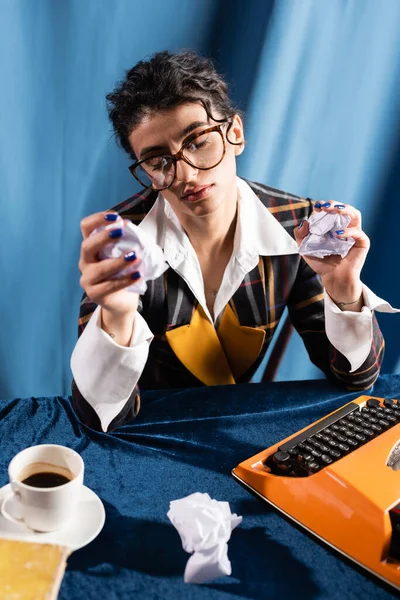 Periodista molesto y agotado sosteniendo papel arrugado cerca de la máquina de escribir sobre fondo azul - foto de stock