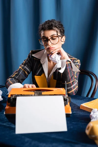 Sad newswoman sitting near vintage typewriter with empty paper sheet on blue background — Stock Photo