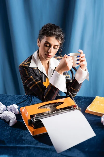 Журналист в стиле ретро с чашкой кофе, сидящей рядом с пишущей машинкой и смятой бумагой на синем фоне — стоковое фото