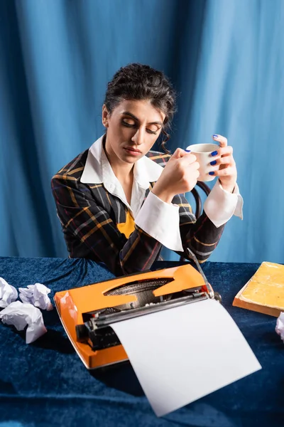 Periodista de estilo retro con taza de café sentado cerca de la máquina de escribir y papel arrugado sobre fondo azul - foto de stock