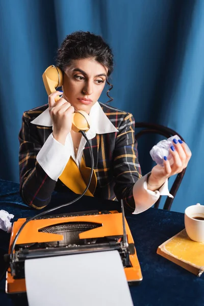 Newswoman pensativo com papel amassado falando no telefone perto da máquina de escrever no fundo azul — Fotografia de Stock