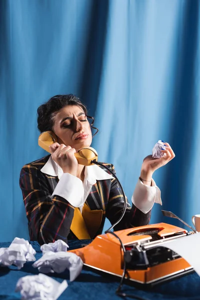 Mujer molesta sosteniendo papel arrugado mientras habla por teléfono cerca de la máquina de escribir sobre fondo azul - foto de stock