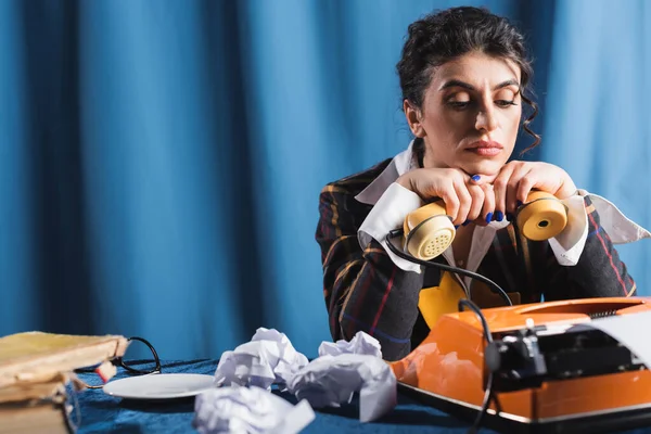 Upset newswoman sitting with handset near vintage typewriter and crumpled paper on blue background — Stock Photo