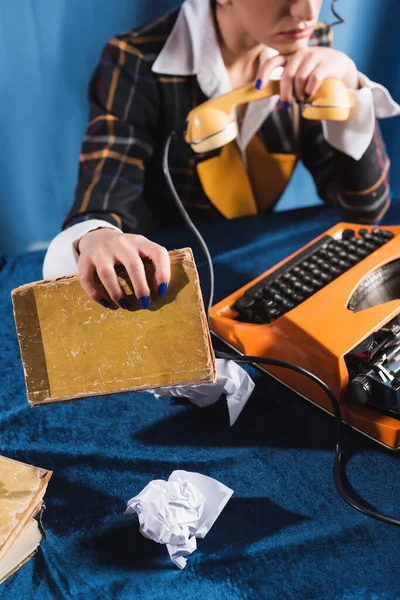 Vista cortada de newswoman segurando livro e fone de ouvido perto de máquina de escrever vintage na toalha de mesa de veludo azul — Fotografia de Stock