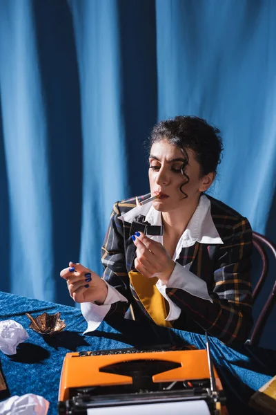 Mujer en estilo vintage chaqueta iluminación cigarrillo cerca de máquina de escribir sobre fondo azul con drapeado - foto de stock