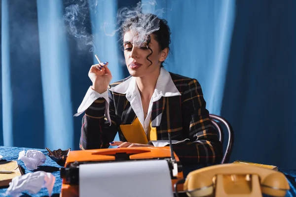 Mujer elegante en ropa de estilo vintage fumar cerca de máquina de escribir y papel arrugado sobre fondo azul - foto de stock