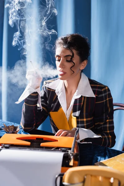 Estilo vintage newswoman mecanografía en la máquina de escribir mientras se fuma sobre fondo azul - foto de stock