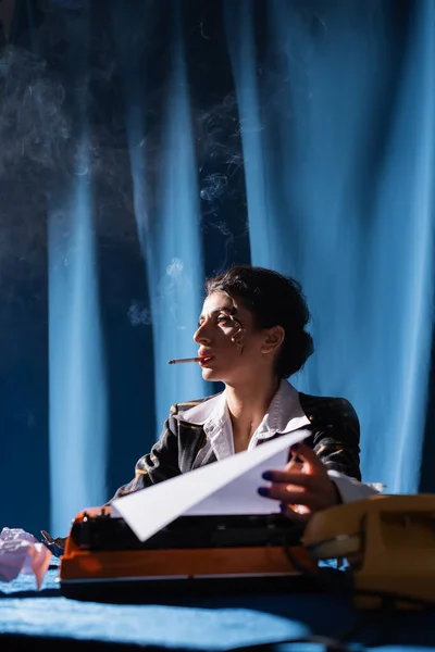 Stylish woman holding paper near blurred typewriter while smoking on background with blue drapery — Stock Photo