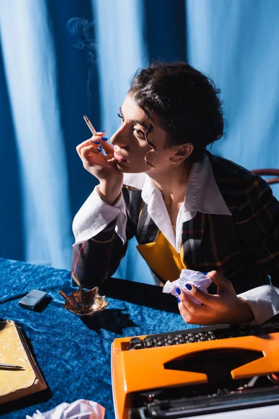 Thoughtful journalist with cigarette and crumpled paper sitting near vintage typewriter on blue background — Stock Photo