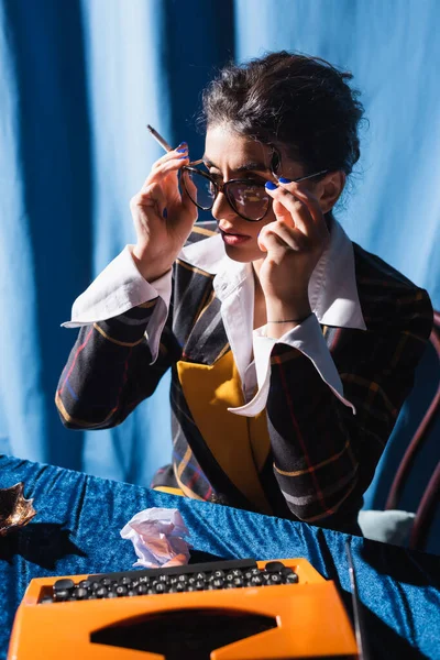 Frau im Retro-Stil mit Zigarettenbrille in der Nähe der Schreibmaschine auf blauem Hintergrund — Stockfoto