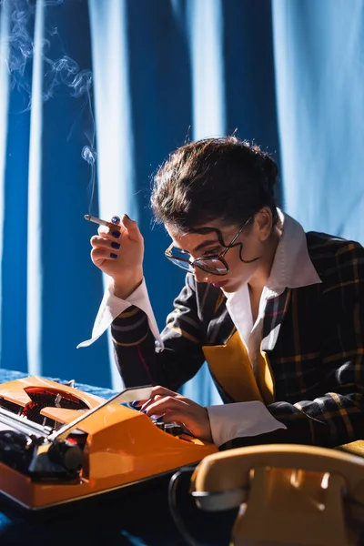 Stylish newswoman in eyeglasses holding cigarette while working at typewriter on blue background — Stock Photo