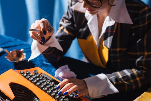 Vista recortada de la mujer borrosa con la escritura de cigarrillos en la máquina de escribir vintage sobre fondo azul - foto de stock