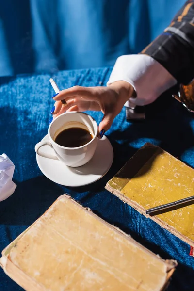 Vue partielle de la femme avec cigarette près de tasse de café et livres vintage sur nappe en velours bleu — Photo de stock