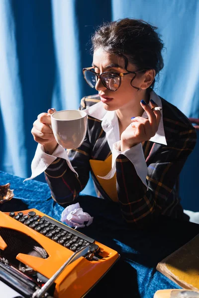 Stile vintage newswoman in occhiali da vista fumare e bere caffè vicino alla macchina da scrivere su sfondo blu — Foto stock