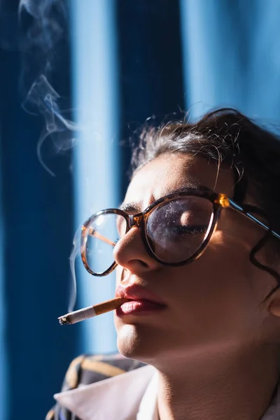 Close up view of woman in vintage eyeglasses smoking with closed eyes on blue background — Stock Photo