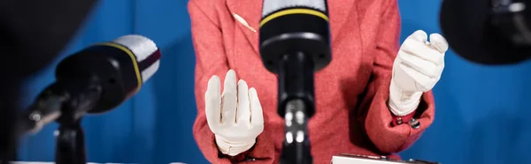 Vue recadrée d'une femme en gants blancs gesticulant près de microphones flous sur fond bleu, bannière — Photo de stock