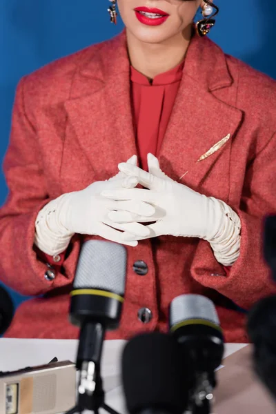 Partial view of woman in white gloves sitting with clenched hands near blurred microphones on blue background — Stock Photo