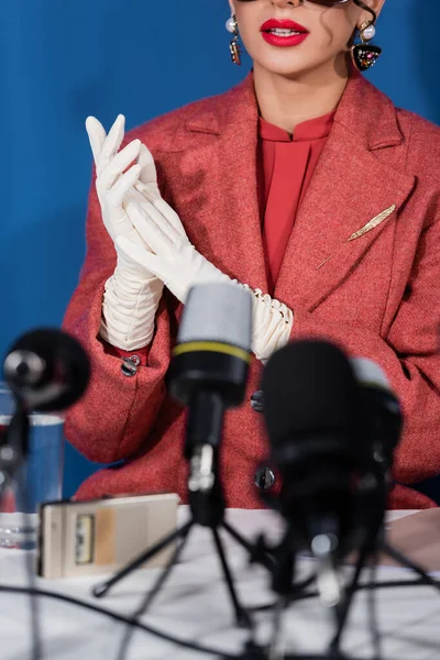 Partial view of stylish woman in red blazer and white gloves near blurred microphones on blue background — Stock Photo