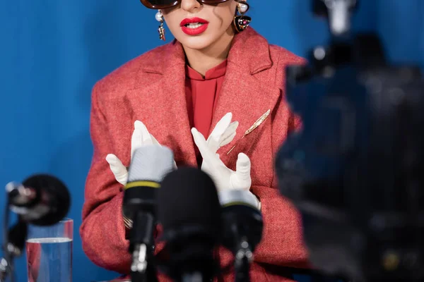 Blurred microphones near cropped woman in white gloves giving interview on blue background — Stock Photo
