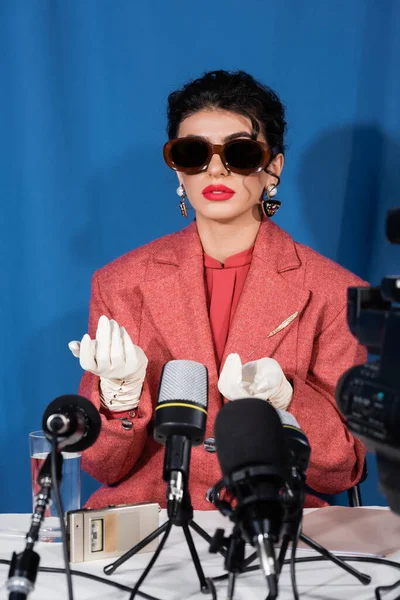 Young woman in vintage sunglasses gesturing during interview on blue background — Stock Photo