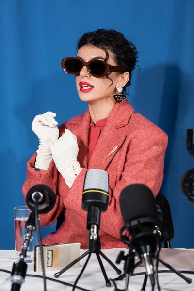 Mujer de estilo vintage en gafas de sol y guantes blancos hablando durante la entrevista sobre fondo azul - foto de stock