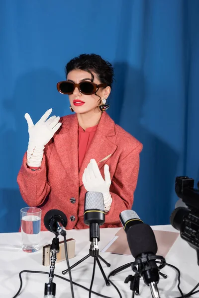 Stylische Frau mit Vintage-Sonnenbrille und weißen Handschuhen gestikuliert beim Interview auf blauem Hintergrund — Stockfoto