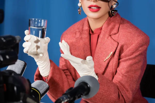 Vue partielle de femme de style rétro avec verre d'eau donnant interview sur fond bleu — Photo de stock