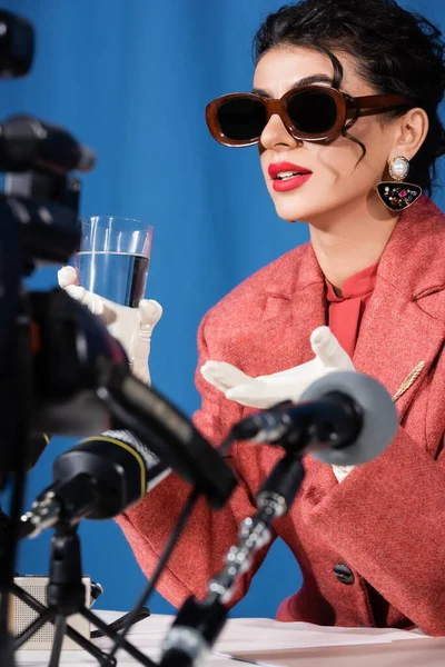 Blurred microphones near retro style woman holding glass of water during interview of blue background — Stock Photo