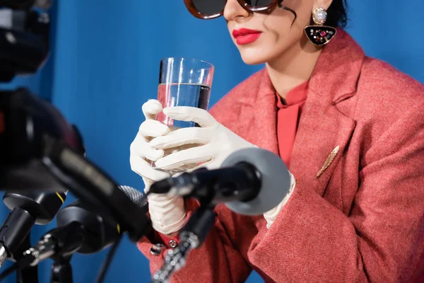 Частичный вид женщины в белых перчатках, держащей стакан воды возле размытых микрофонов на синем фоне — стоковое фото
