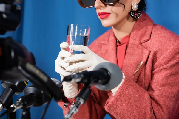 Vista parcial de la mujer de estilo retro en guantes blancos sosteniendo un vaso de agua cerca de micrófonos borrosos sobre fondo azul - foto de stock