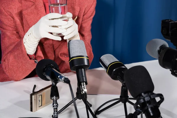 Cropped view of woman in white gloves holding glass of water near microphones and voice recorder on blue background — Stock Photo