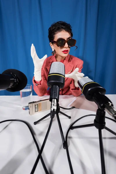 Micrófonos cerca de la mujer de estilo vintage en gafas de sol gesto durante la entrevista sobre fondo azul - foto de stock