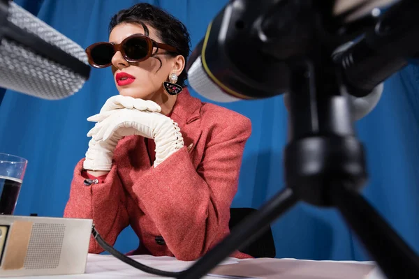 Micrófonos borrosos cerca de la mujer con estilo en gafas de sol vintage y guantes blancos sobre fondo azul - foto de stock