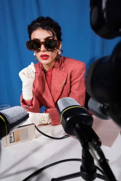 Femme de style rétro dans des lunettes de soleil parler près de microphones flous sur fond bleu — Photo de stock