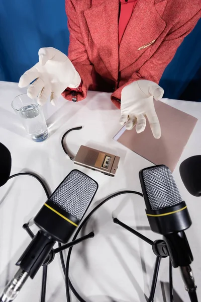 Vista superior de micrófonos, grabadora de voz y vaso de agua cerca de mujer recortada en guantes blancos sobre fondo azul - foto de stock