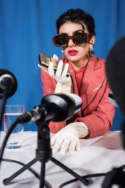 Micrófonos borrosos cerca de la mujer de estilo retro que sostiene la grabadora de voz durante la entrevista sobre fondo azul - foto de stock